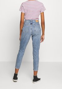 Džínové kalhoty s vysokým pasem v modré barvě, rovného střihu, s dvěma zadními kapsami a nášivkou na pase. Nosí se s pruhovanou košilí a černými teniskami.