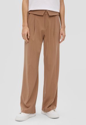 Personne portant un pantalon marron taille haute et jambes larges, associé à des baskets blanches et un haut blanc rentré, sur un fond uni.