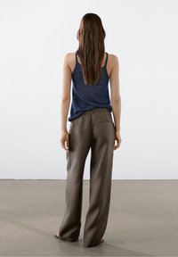 Vrouw die blootsvoets staat, met haar rug naar de kijker, in een mouwloos marineblauw topje en losse bruine broek, tegen een effen lichte achtergrond.