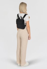Svart ryggsäck med justerbara remmar, slätt tyg, framficka och logodetalj; bärs över en vit skjorta och beige ribbade byxor.