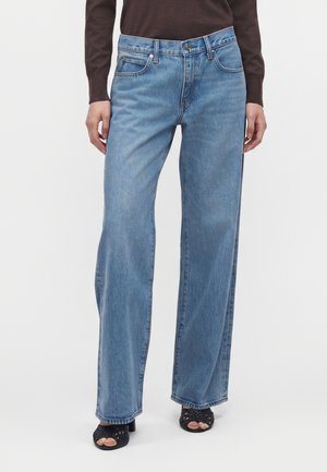 Person iført lyseblå vidde jeans og sorte åbne hælet sandaler, stående mod en hvid baggrund.