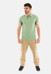 Polo shirt de manga corta verde claro con cuello clásico, combinado con pantalones cargo beiges y zapatillas oscuras, sobre un fondo blanco.
