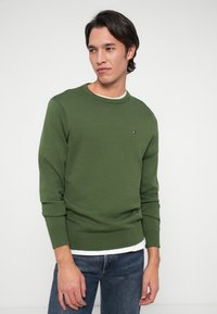 Πράσινο πλεκτό πουλόβερ με στρογγυλή λαιμόκοψη, ραμμένες μανσέτες και στρίφωμα. Διαθέτει ένα μικρό λογότυπο στο στήθος. Συνδυάζεται με μπλε τζιν παντελόνι.