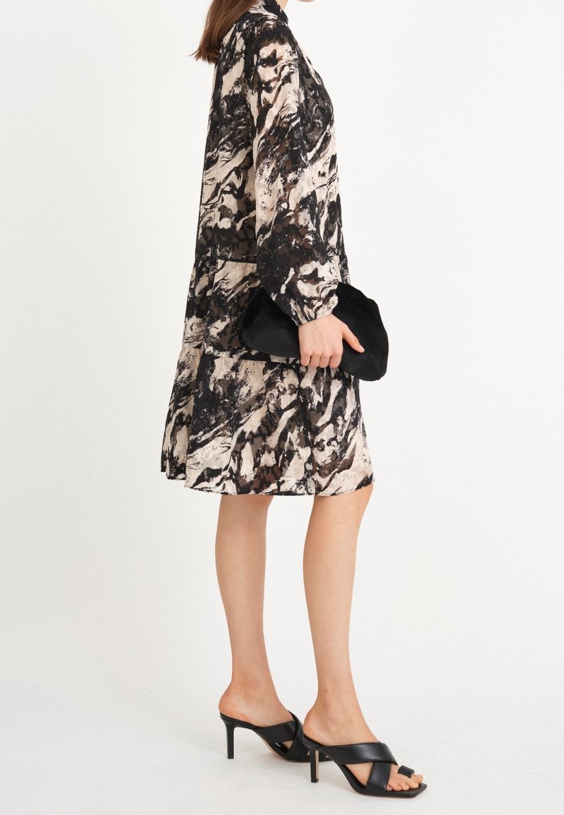 Robe imprimée beige, noire et marron, avec des manches longues et une coupe ample. Portée avec des sandales à talons hauts noires et une pochette noire.