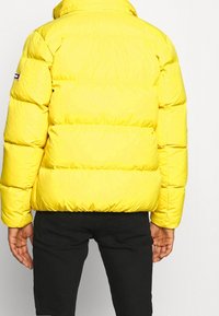 Žlutá péřová bunda s prošívaným designem. Má vysoký límec a viditeln�é švy. Pod bundou jsou nošeny černé kalhoty.