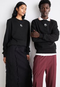 Sweatshirts noirs avec logo sur la poitrine, associés à une jupe noire à franges et un pantalon bordeaux ; présentation de textures et couleurs variées.