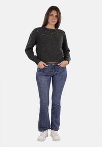 Maglione grigio lavorato a maglia con decorazioni sul collo, abbinato a jeans blu a zampa d'elefante e calzature bianche, esposto su uno sfondo bianco.