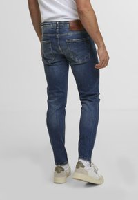 Jeans de denim albastru închis, cu un croi slim, buzunare spate și o etichetă din piele maro. Purtați cu teniși albi și gri, având un aspect ușor uzat.