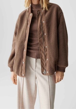 Brązowa kurtka polarowa z zapięciem na zatrzaski i zamek błyskawiczny, ściągaczami na mankietach i gładkimi akcentami materiału z przodu. Noszona na brązowym swetrze z dzianiny.