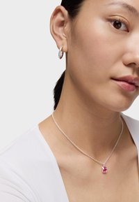 Collana a catena d'argento con un pendente rosa a forma di orsetto. Il modello indossa una semplice maglietta bianca e orecchini a cerchio. Stile pulito e minimalista.