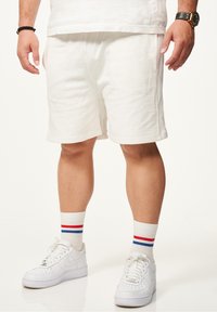 Weiße Baumwollshorts mit relaxed Fit, kombiniert mit weißen Sneakern und Socken mit roten und blauen Streifen. Einfaches Design, legere Kleidung.