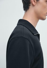 Zwarte polo met kraag en ritssluiting, vervaardigd van een textuurstof met een gladde afwerking. Zijaanzicht benadrukt de kraag en schouderdetails.