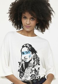 Weißes Oversized-T-Shirt mit einem schwarz-blauen Grafikdesign einer Frau mit Federn und Rosen. Weicher Stoff mit entspannter Passform und überschnittenen Ärmeln.