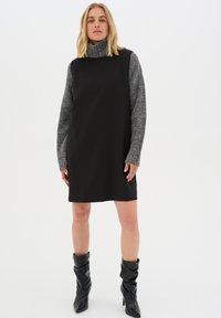 Schwarzes ärmelloses Kleid mit hohem Kragen, ergänzt durch graue strukturierte lange Ärmel. Trägt man mit schwarzen knielangen Lederstiefeln.