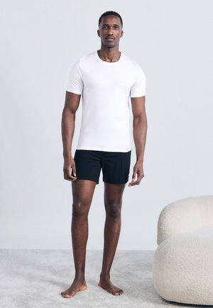 Hombre de pie descalzo sobre una alfombra, vistiendo una camiseta blanca de manga corta y pantalones cortos negros, contra un fondo simple de color gris claro.