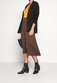 Schwarzer Blazer, orange Bluse, brauner Midirock mit kleinem schwarz-orangen Muster und schwarze Ankle Boots mit Blockabsatz. H�ält eine schwarze Tasche.