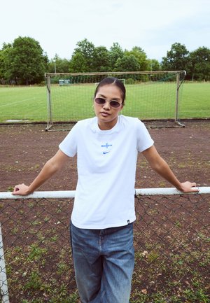 White cotton t-shirt with blue Nike logo, worn with relaxed blue jeans; person stands against a fence on a grassy field, wearing sunglasses.