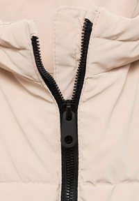 Ljust beige puffer-jacka med en svart dragkedja. Har en dragkedjestängning, vadderad textur och en rund dragkedjehållare.