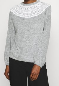 Šedý svetr s dlouhým rukávem, který má bílé krajkové detaily s jedinečnými vzory. Měkká texturovaná tkanina, kulatý výstřih a elastické manžety.