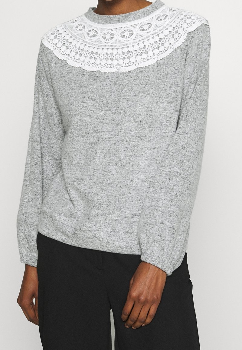 Šedý svetr s dlouhým rukávem, který má bílé krajkové detaily s jedinečnými vzory. Měkká texturovaná tkanina, kulatý výstřih a elastické manžety.