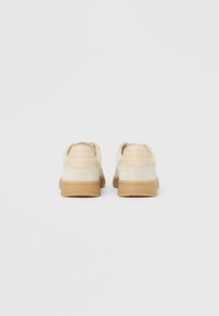 Poros smėlio spalvos zomšinių sportinių batelių su smėlio spalvos paminkšlintais kulnais ir gumos pado padais, matomi iš nugaros, ant paprasto pilko fono.