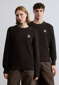 Pulls en maille côtelée brun foncé à manches longues, col rond, avec un petit logo losange blanc sur la poitrine. Associés à un pantalon marron.