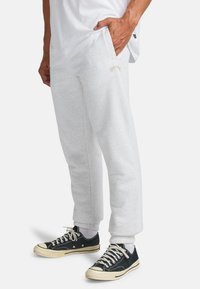 Lichtgrijze trainingsbroek met zijzakken, geribde manchetten en een geborduurd logo in goud. Draag het met zwarte sneakers en witte sokken.