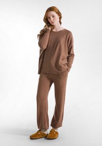 Bruin gebreid loungewear set met een lange mouw top met zijzakken en bijpassende wijde broek, aangevuld met bruine slippers.