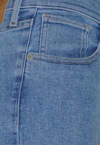 Gros plan d'un jean en denim bleu montrant la poche avant avec rivet, couture et une partie de la boucle de ceinture.