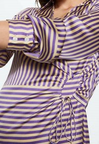 Violetti ja beige raidallinen paita, jossa on napitettu hihansuu, kerättyjä sivuosi yksityiskohtia ja solmittava suljenta, valmistettu sileästä, kevyestä kankaasta.