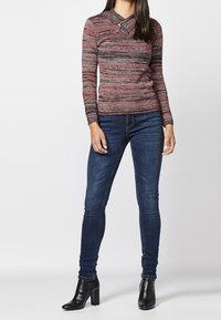 Haut à manches longues rayé rouge et noir avec un léger col montant et un détail zip, associé à un jean skinny bleu foncé et des bottines noires.