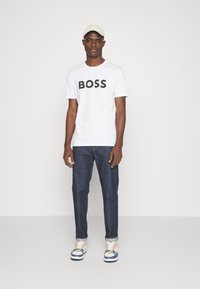 T-shirt en coton blanc avec "BOSS" en lettres noires en gras, associé à un jean en denim bleu et des baskets blanches avec des accents bleu et beige.