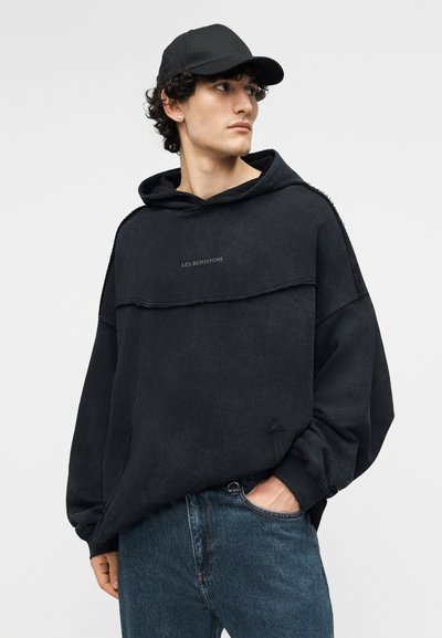 Junger Mann mit schwarzer Kappe und übergroßem schwarzem Hoodie mit dem Schriftzug "LES BENJAMINS", eine Hand in der Tasche seiner blauen Jeans, schaut nach rechts.