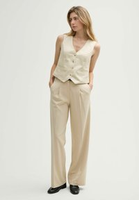 Beige Leinenweste mit V-Ausschnitt, Knopfverschluss und Taschen, kombiniert mit beigen weit geschnittenen Hosen. Das Model steht mit den Händen in den Taschen.