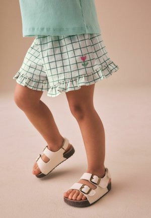 Kind trägt grüne karierte Rüschen-Shorts mit rosa Blumenstickerei und weiße Sandalen mit Schnallenriemen, steht auf hellem Boden.