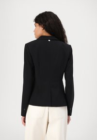 Svarte skreddersydde blazere med spisse lapeller, enkelt bakventil og detalj i sølvknapp nær kragen. Stoffet har en jevn tekstur.