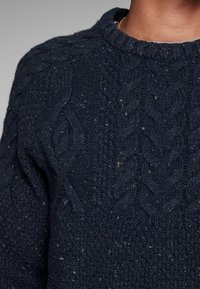 Donkerblauwe kabelgebreide trui met textuurpatronen en een geribbelde halslijn. Zachte stof met subtiele spikkels overal.