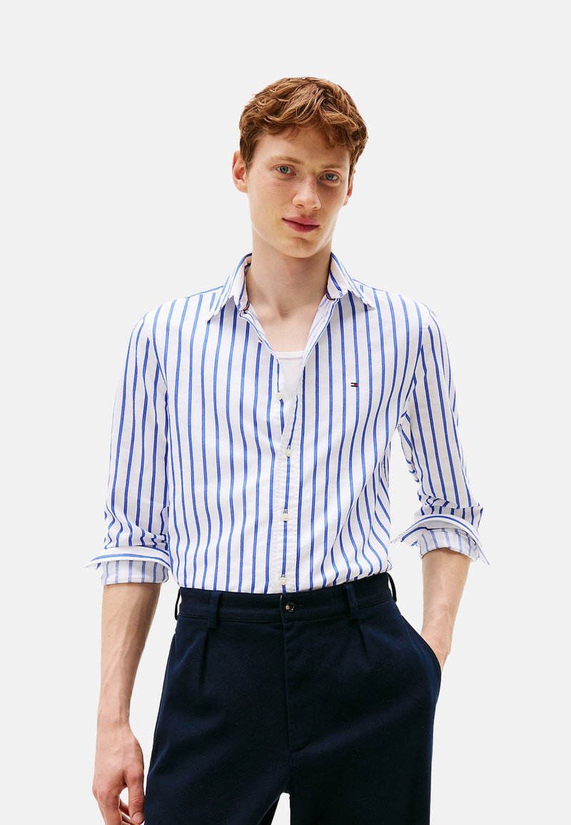 Camicia bianca con righe verticali blu, con colletto e maniche arrotolate. Indossata con pantaloni scuri, che mostrano una texture liscia.