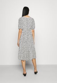 Biała sukienka z czarnym wzorem serc, z krótkimi bufkami, okrągłym dekoltem, marszczoną w talii i półdługą spódnicą w wariancie warstwowym. Czarne sandały na blokowym obcasie.