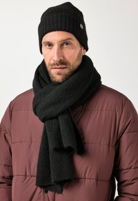 Schwarze Mütze und Schal aus weichem Strickmaterial. Die Mütze hat eine gerippte Struktur; der Schal ist breit und fällt leicht. Beide Artikel sind schlicht und ohne Verzierungen.