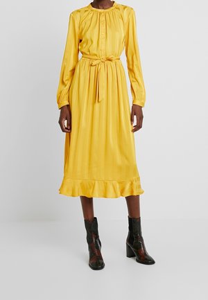 Femme portant une robe midi jaune à manches longues avec une ceinture à la taille et un ourlet à volants, associée à des bottes noires mi-mollet à talons.