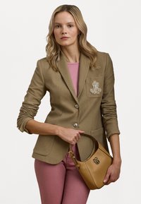 Blazer beige su misura con bottoni argentati e dettagli ricamati sulla tasca, abbinato a un top rosa e pantaloni rosa, mentre tiene una borsa color nocciola.
