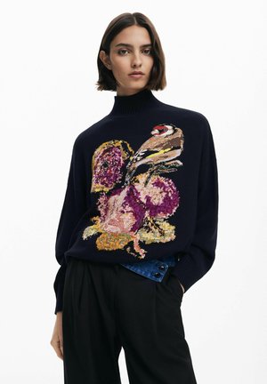 Donna che indossa un maglione blu navy con collo alto decorato con ricami colorati di uccelli e fiori, abbinato a pantaloni neri e un dettaglio di cintura in denim.