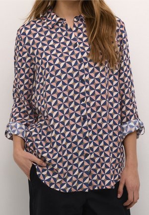 Femme portant une chemise à manches longues boutonnée avec un motif circulaire géométrique rose, marine et beige et des poignets retroussés.