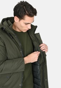 Giacca imbottita color verde oliva con cappuccio, chiusura con zip e bottoni a pressione. Presenta un maglione lavorato a maglia testurizzata sotto e una tasca interna.
