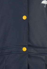 Jūras spēku ūdensizturīga lietus jaka ar dzelteniem pogām, gludu tekstūru un baltu lietussarga grafiku krūšu daļā.