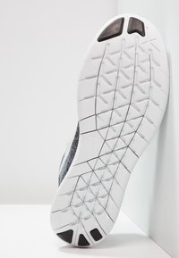 Suola di scarpa sportiva bianca con tacchetti neri e motivo a freccia, inclinata contro un muro e una superficie bianchi.
