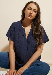 Blusa blu navy con maniche corte, strisce verticali testurizzate e scollatura a V. Abbinata a jeans blu e con uno sfondo chiaro.