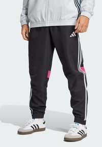Schwarze Jogginghosen mit pinken und weißen Streifen, elastischen Bündchen und einem Logo am Oberschenkel. Kombiniert mit weißen und schwarzen Sportschuhen.