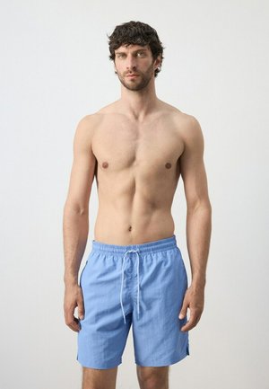 Uomo senza maglietta con capelli corti e scuri, indossa pantaloncini da bagno azzurri, in piedi contro uno sfondo chiaro e uniforme.